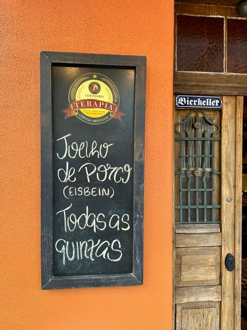 Quadro-negro na fachada anunciando joelho de porco (Eisbein) todas as quintas, ao lado da porta com a placa Bierkeller